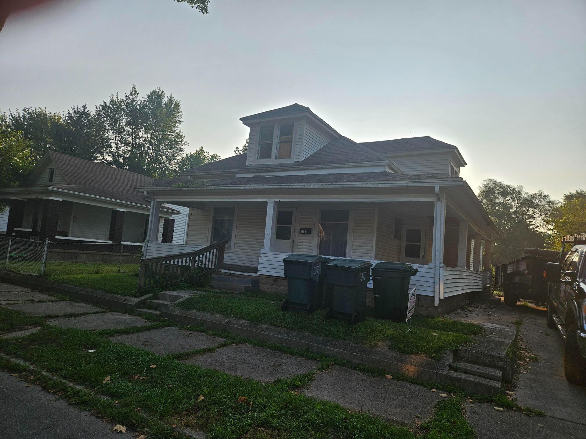 Dilapidated white house with porch and trash cans on a city street.