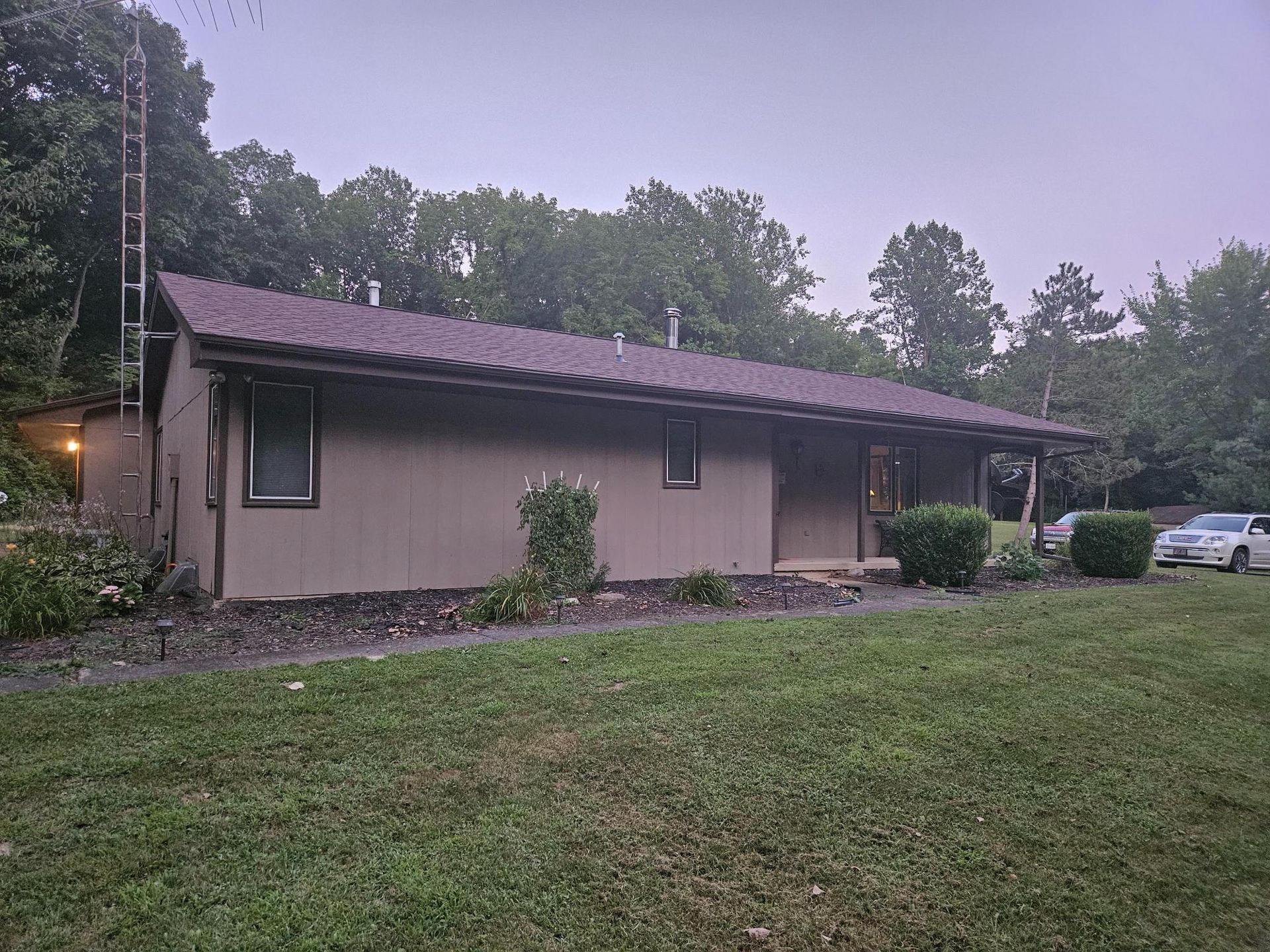 Tan house with brown roof, surrounded by green grass and trees. Cloudy sky.