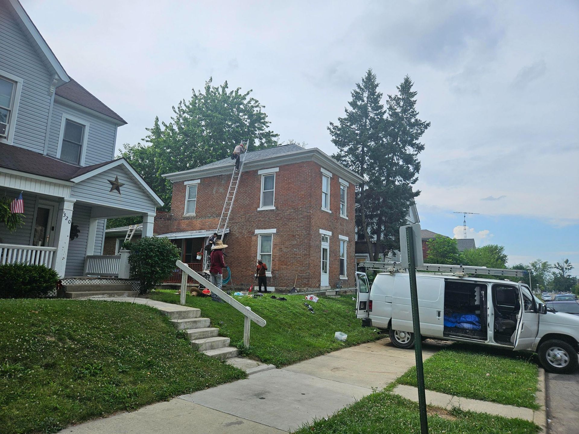 Two-story brick house with workers on ladder; white van parked on the side. Green lawn and cloudy sky.