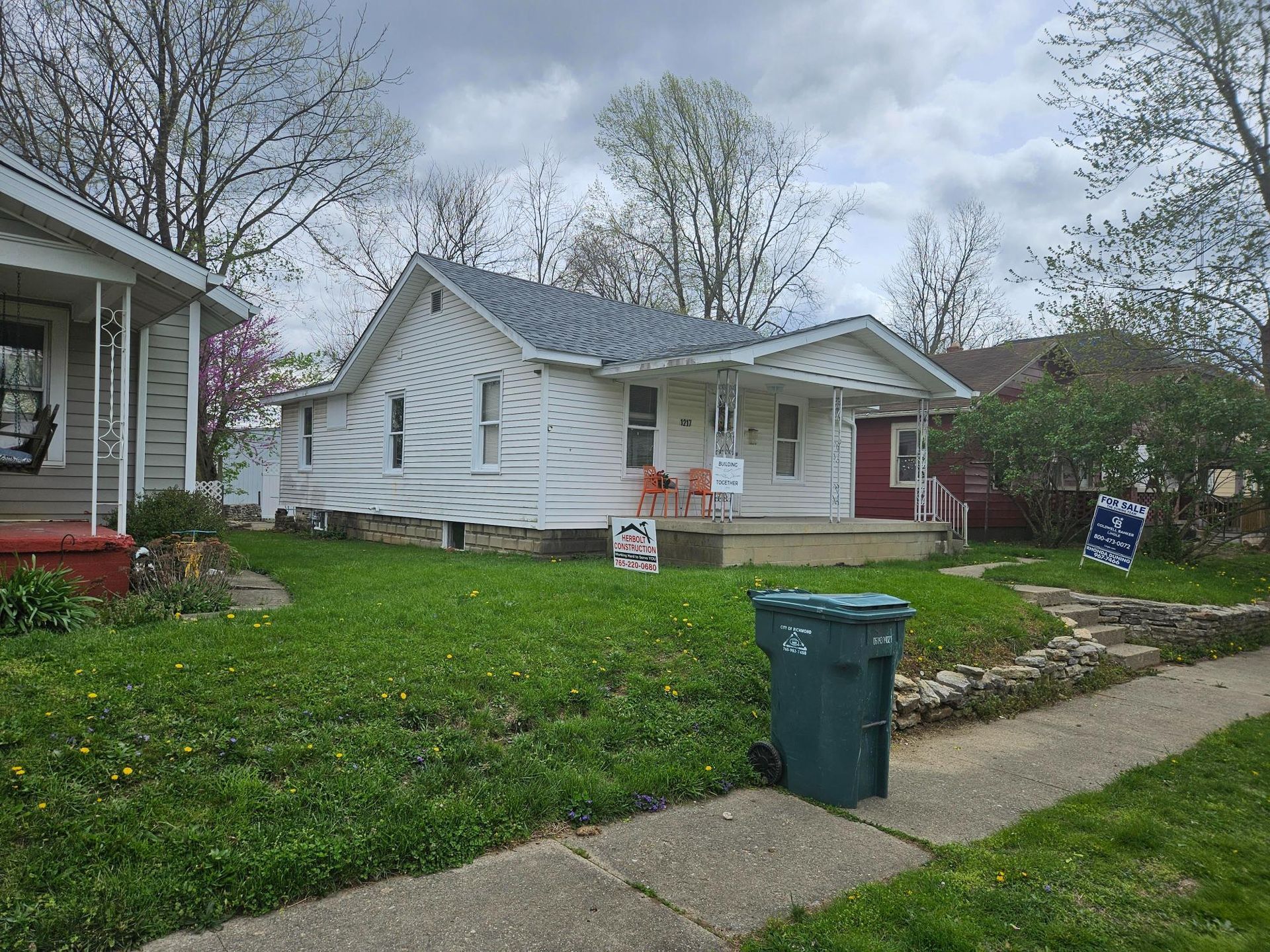 White house with a small porch and front lawn. A green trash can sits in front.