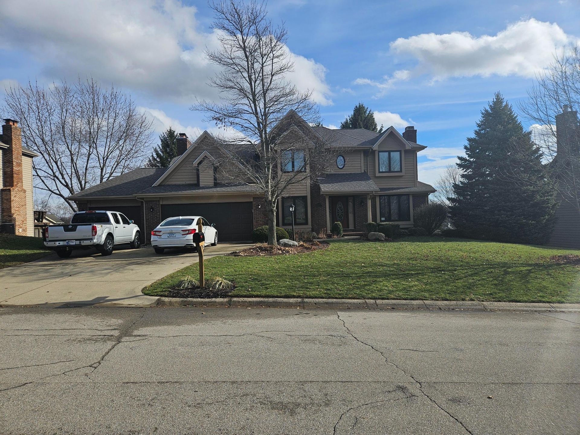 Two-story beige house with a black roof, driveway, and vehicles parked out front; blue sky and clouds above.