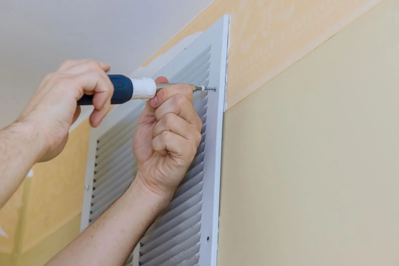 Hands using a screwdriver to install a vent cover on a beige wall