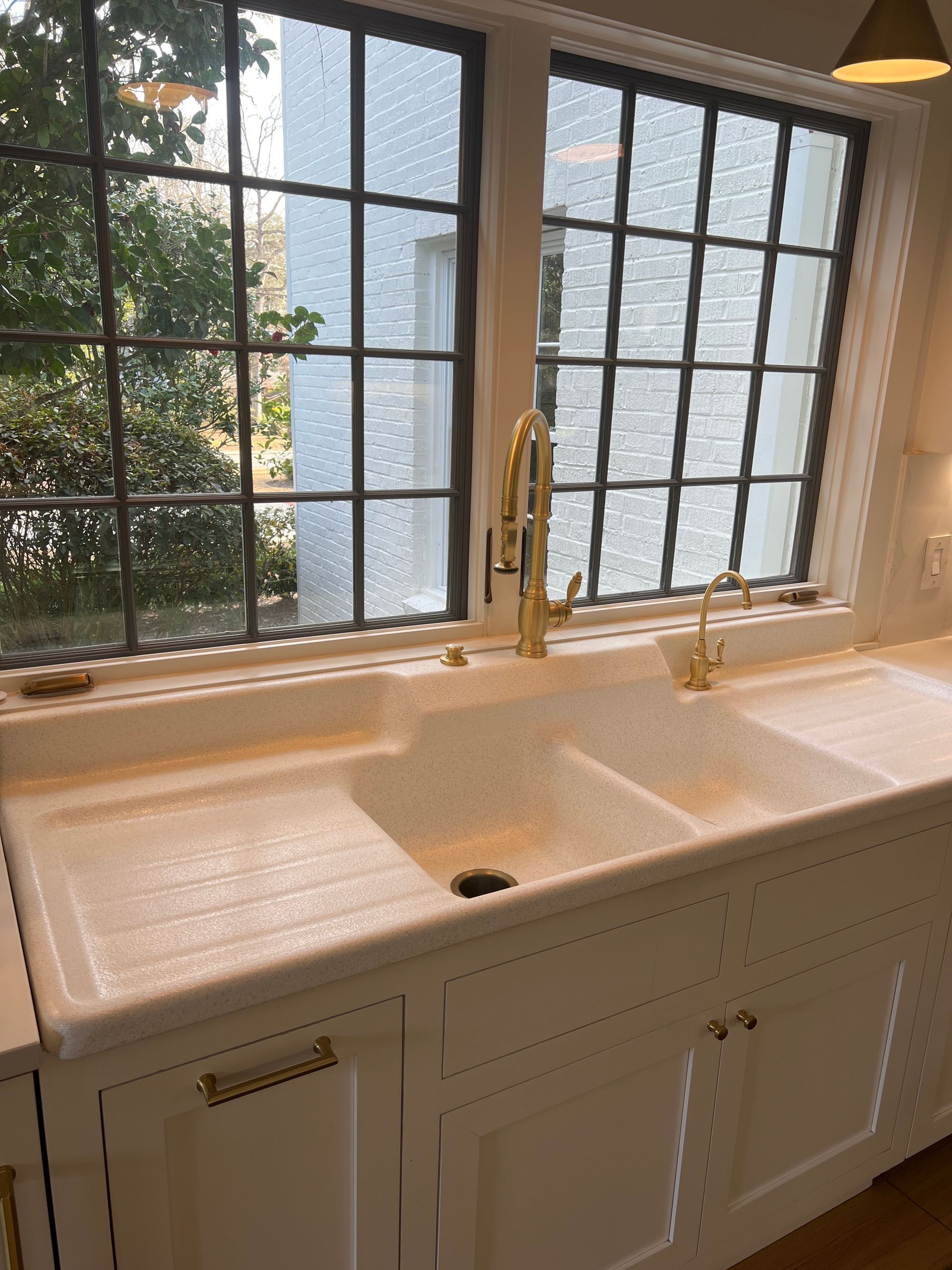 White kitchen sink and gold faucet beneath a window with frosted glass panels and greenery outside