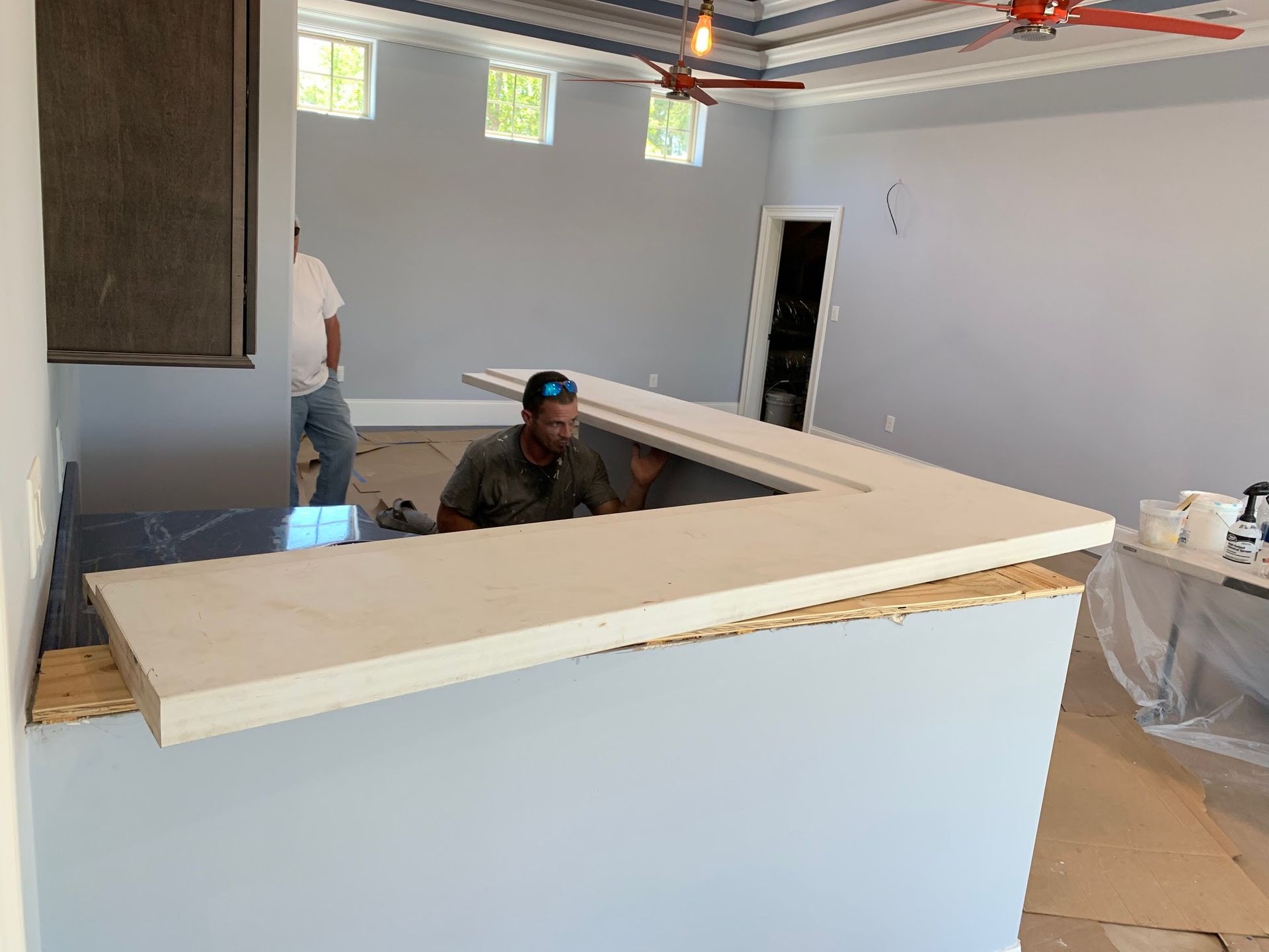 Partially built kitchen island in a bright room, with two workers and a ceiling fan above.