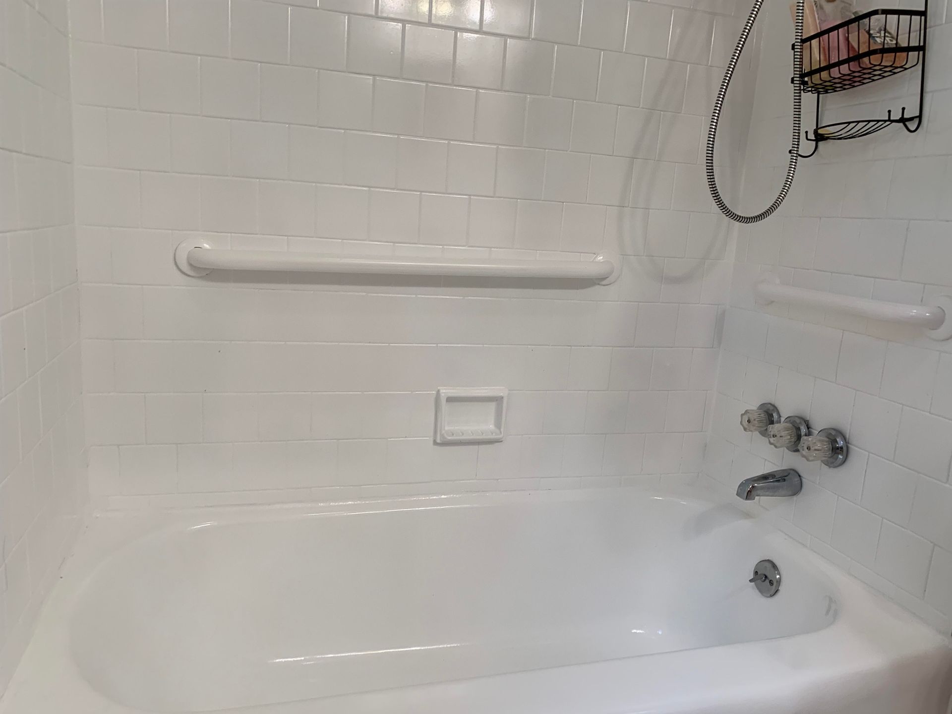 White tiled bathtub and shower with grab bars and wall-mounted controls in a bathroom.