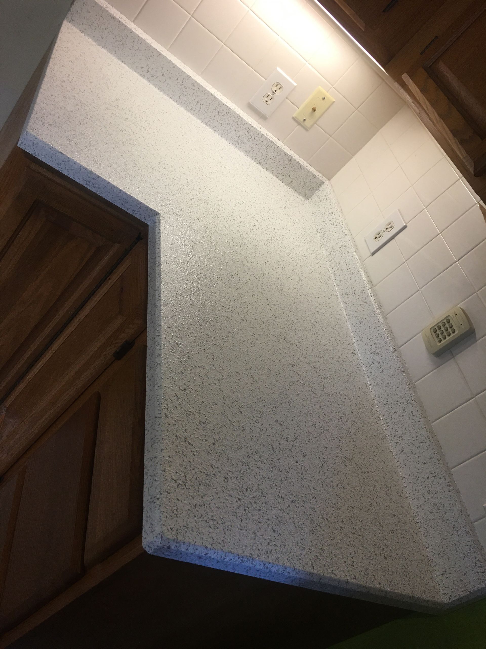 Tilted view of a kitchen corner with speckled countertop, white tile wall, and wooden cabinet