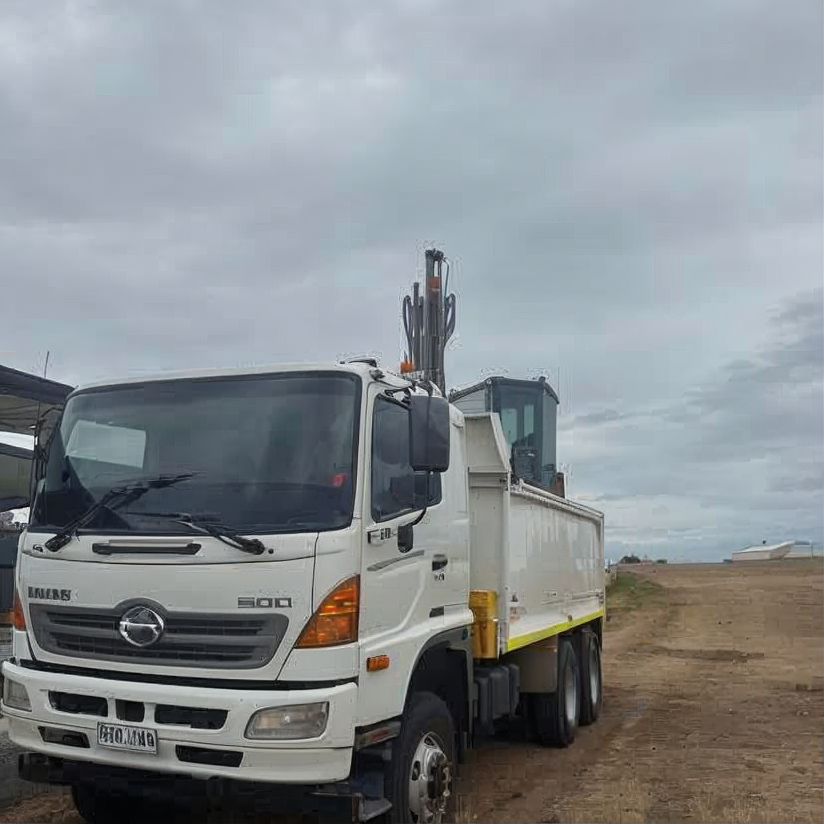 White dump truck on a dirt road under a cloudy sky, with a boom mounted behind the cab.