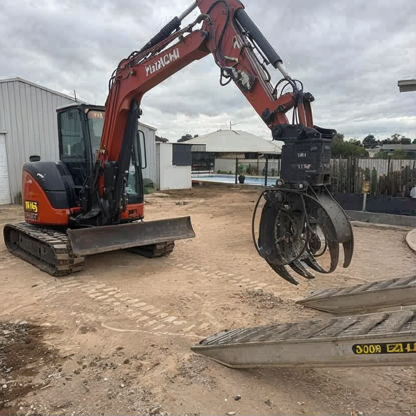 Orange excavator with grapple attachment parked on a dirt lot near buildings