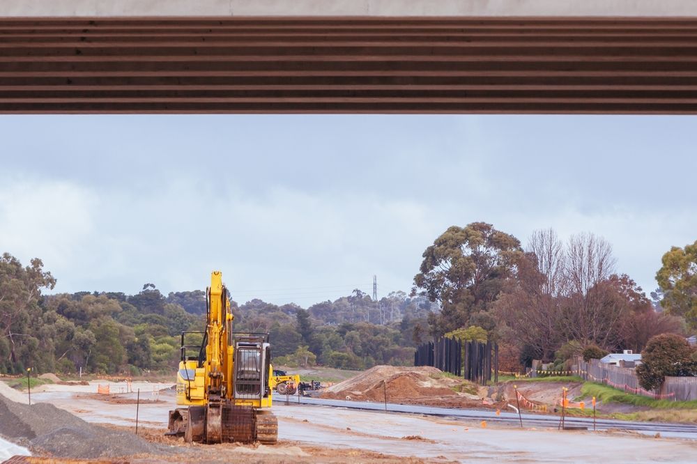 Yellow excavator on a construction site with muddy ground, trees, and a road under overcast sky