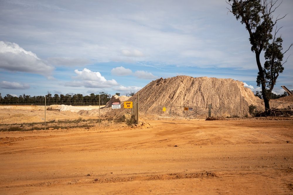 Dry dirt road beside a large sand mound under a cloudy sky, with a lone tree and small signpost.