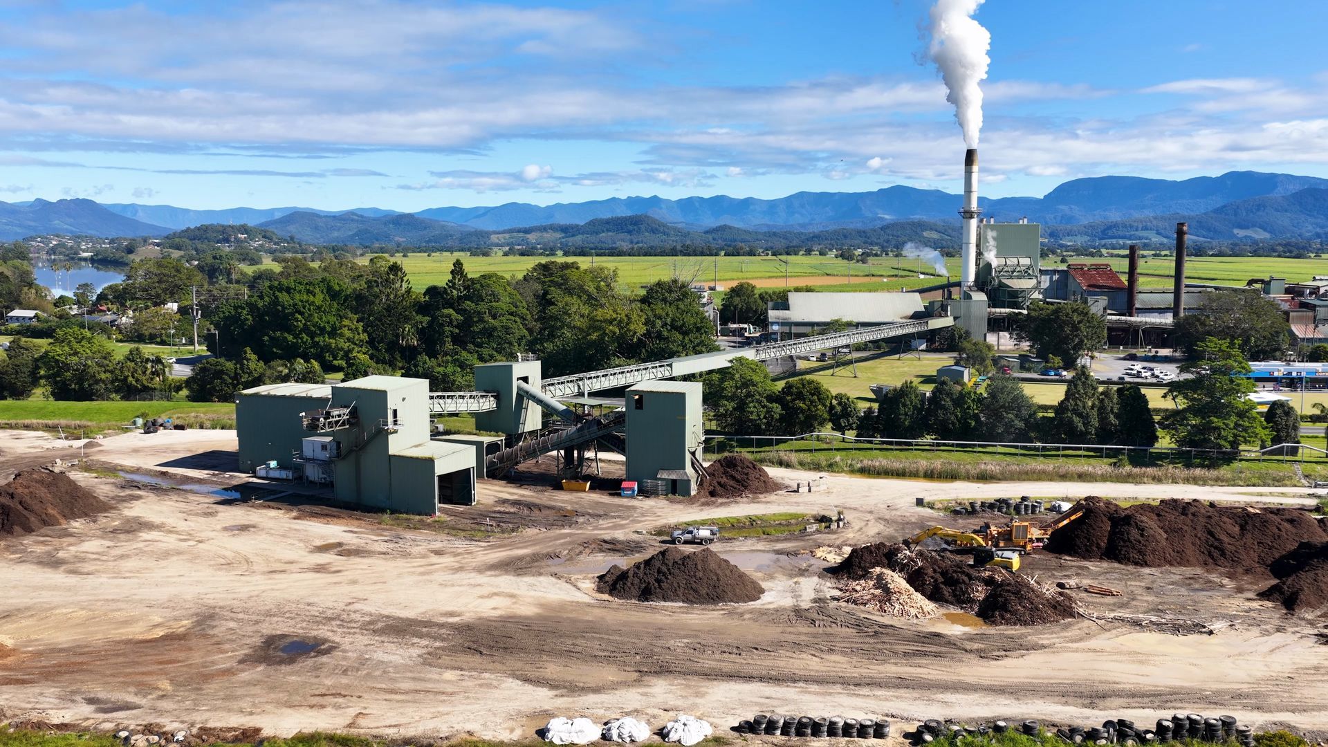 Industrial plant with smokestack in a rural valley, set against mountains and a blue sky.