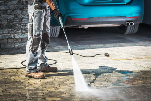 Person pressure washing a driveway beside a blue car and stone wall.
