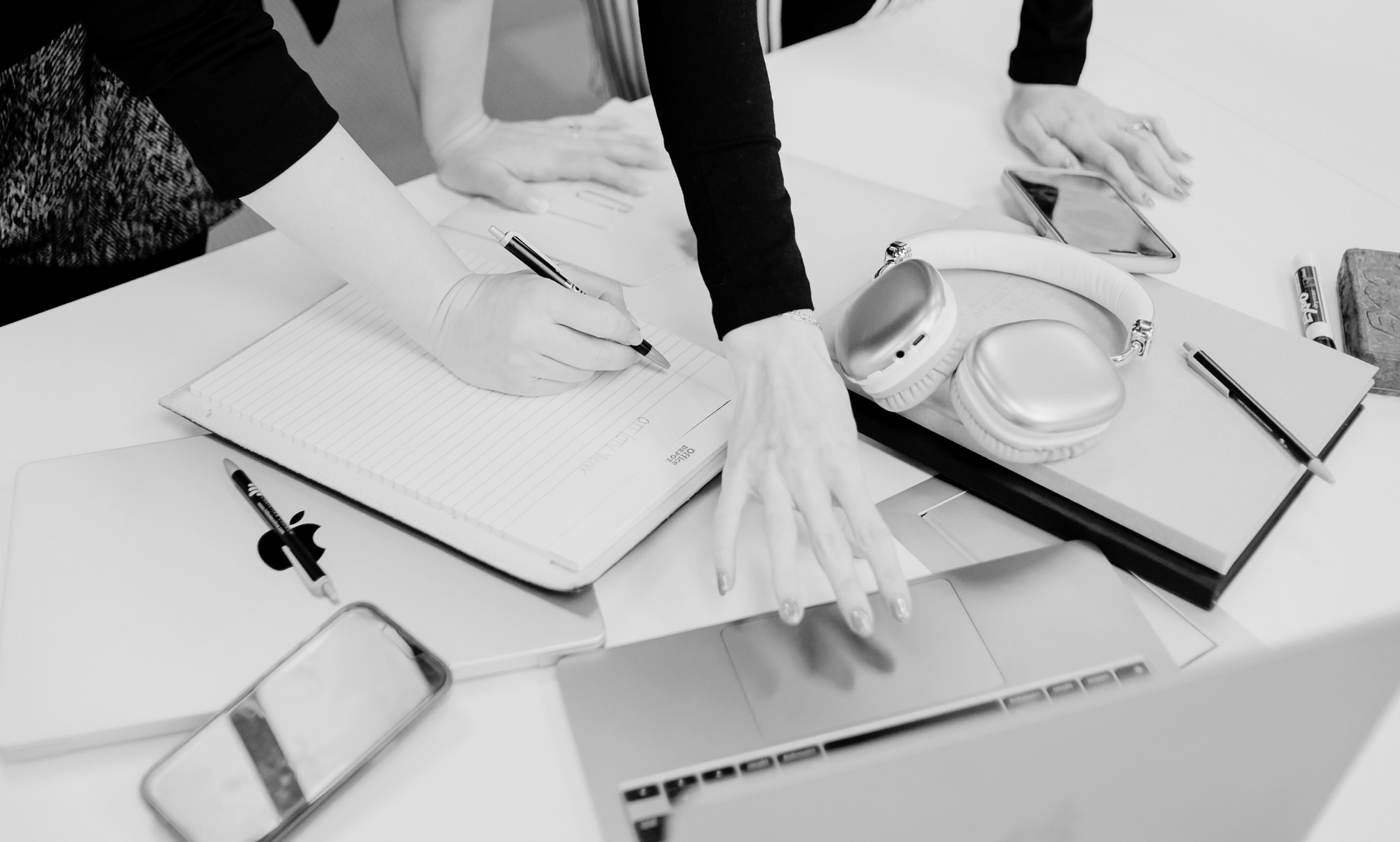 Hands writing on paper, with laptops, headphones, and a phone on a white table.