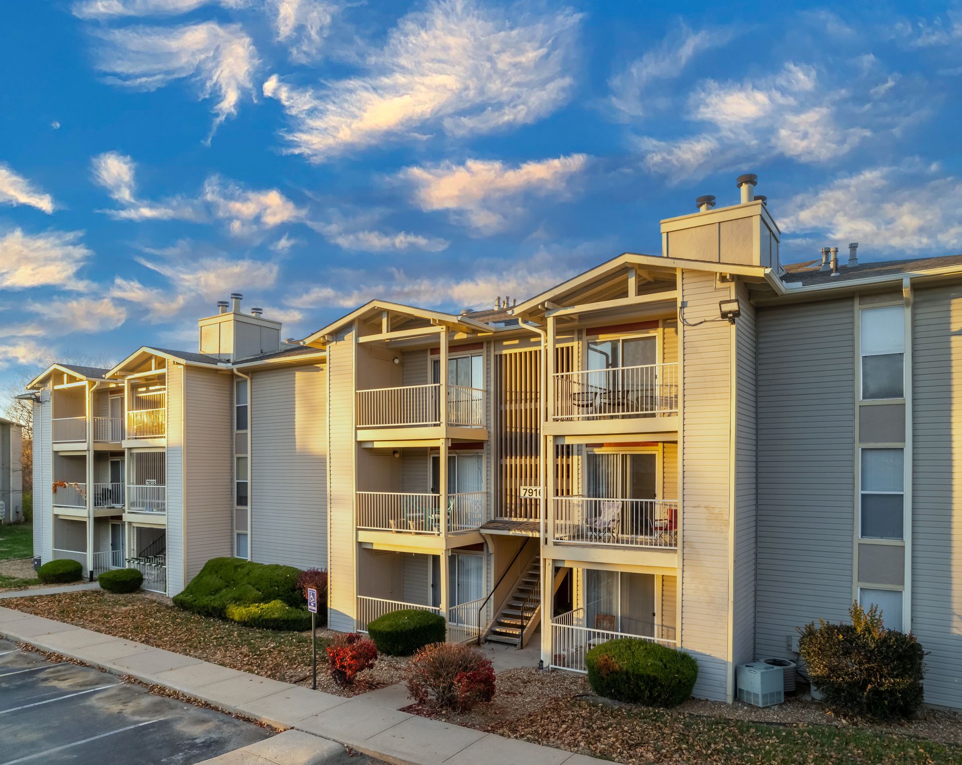 Apartment building exterior, light beige with balconies and stairwells. Blue sky with clouds overhead.