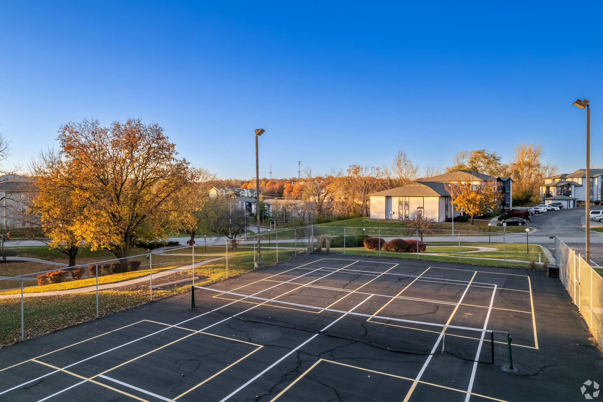 Tennis court with lines, netting, and lights under a blue sky, trees in the background, residential area.