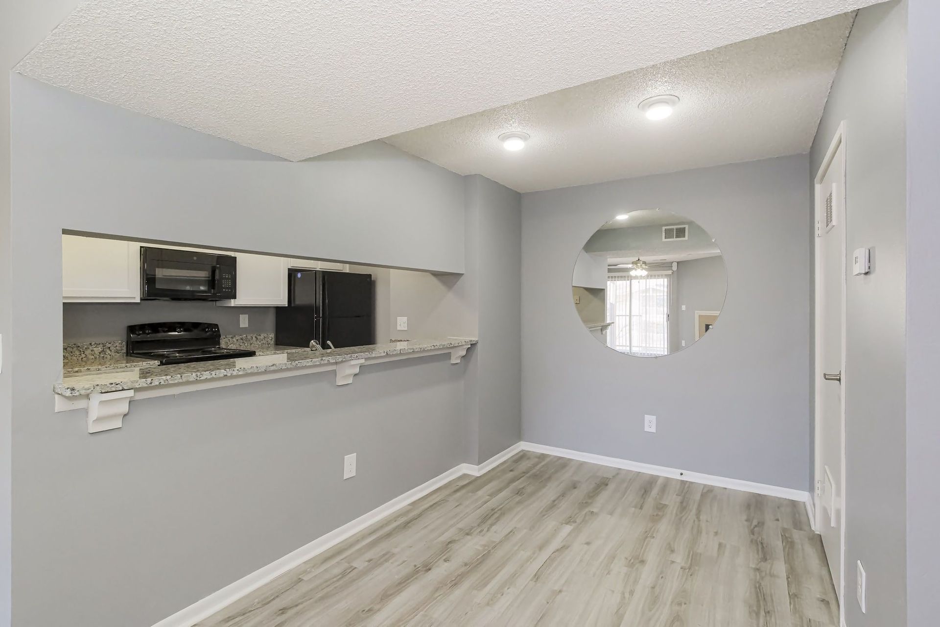 Interior of a dining area with a pass-through to a kitchen; gray walls, wood-look flooring, and a round mirror.