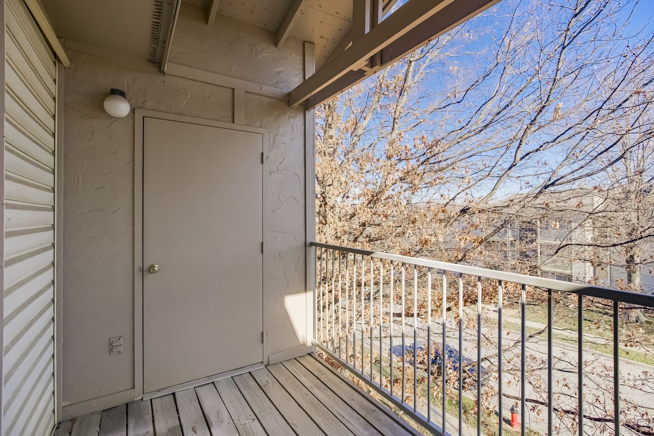 Balcony with beige door, railing, and wooden floor. Bare tree branches in background. Blue sky visible.
