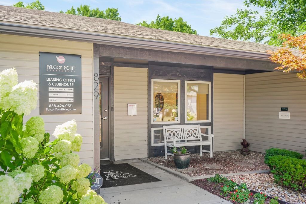 Exterior of a light beige building with a sign and a bench. White flowers are in front.