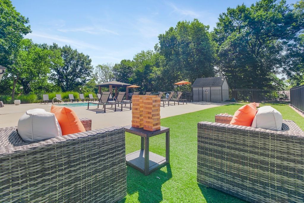Two wicker chairs with orange pillows face a Jenga game on a small table near a pool.