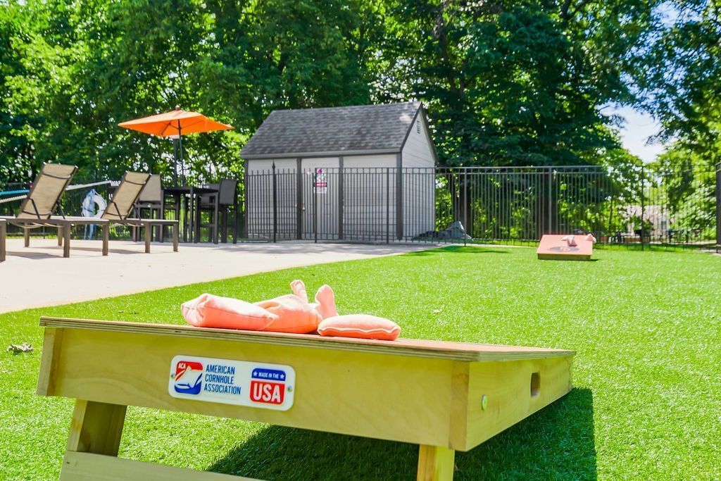 Cornhole set on green turf with building, seating, and trees in background.