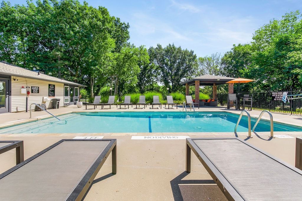 Pool area with lounge chairs, blue water, and trees under a clear sky.