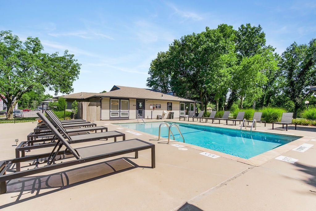 Pool with lounge chairs and building, under blue sky with trees.