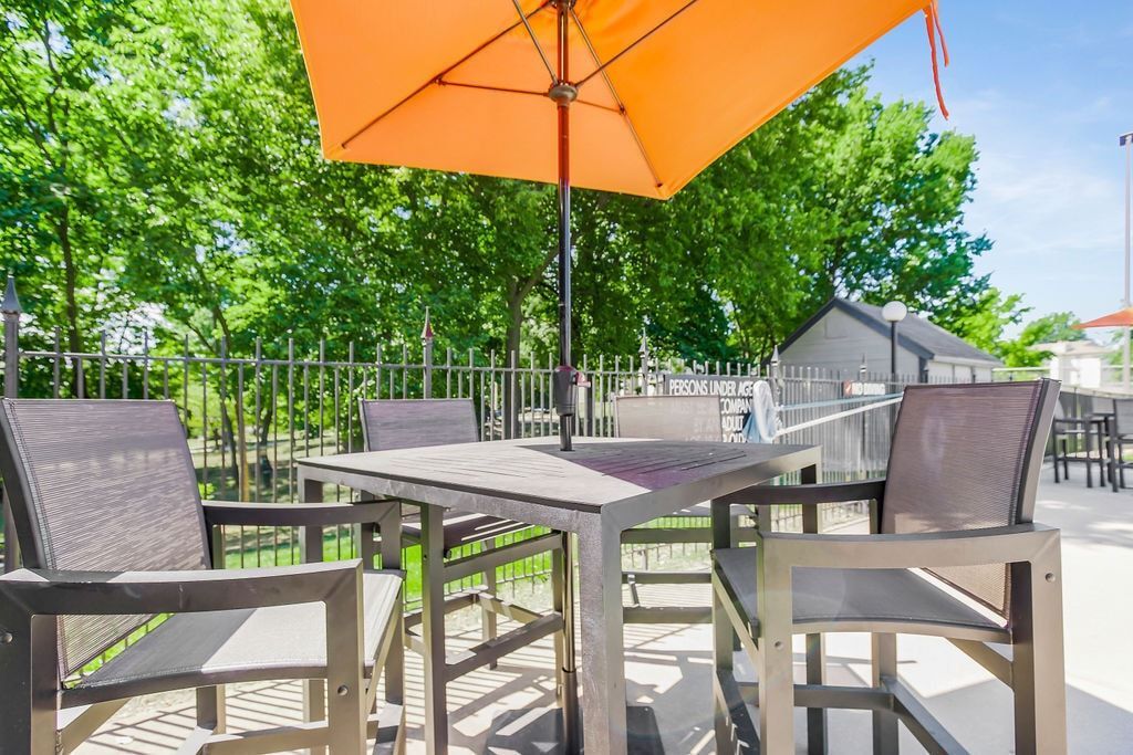 Patio seating with table, chairs, and orange umbrella; trees and fence in background.
