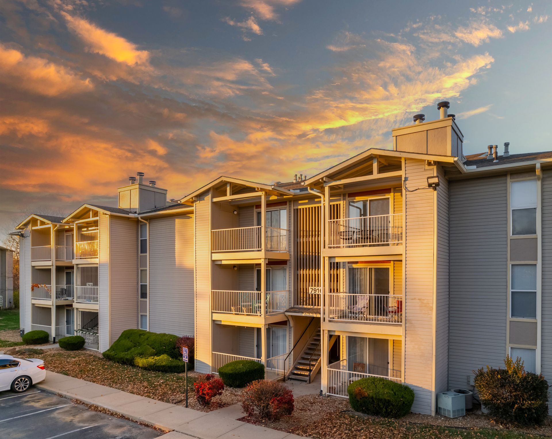 Apartment building exterior at sunset; beige siding, balconies, colorful sky, car in parking area.