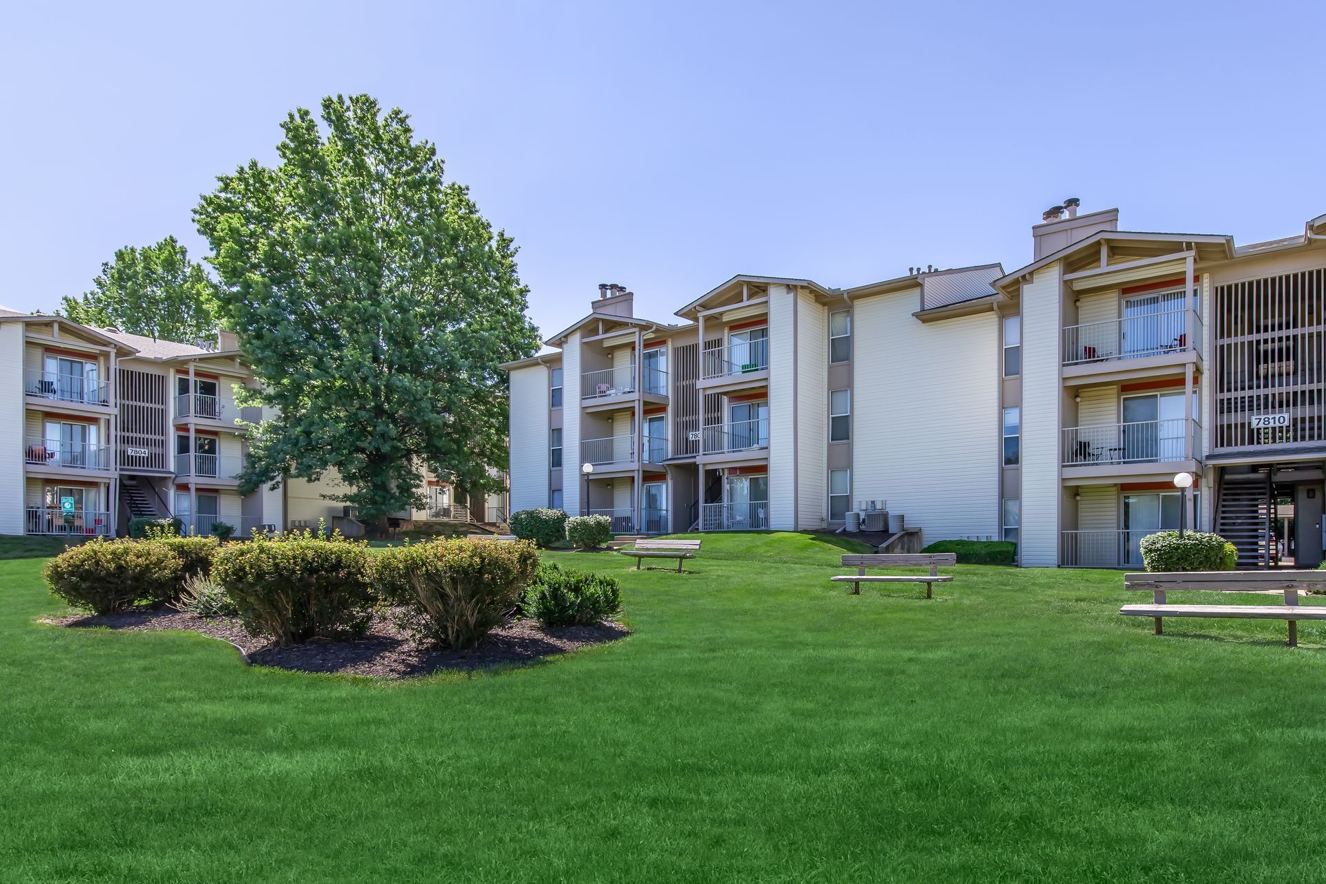 Apartment complex with green grass, bushes, and benches on a sunny day.