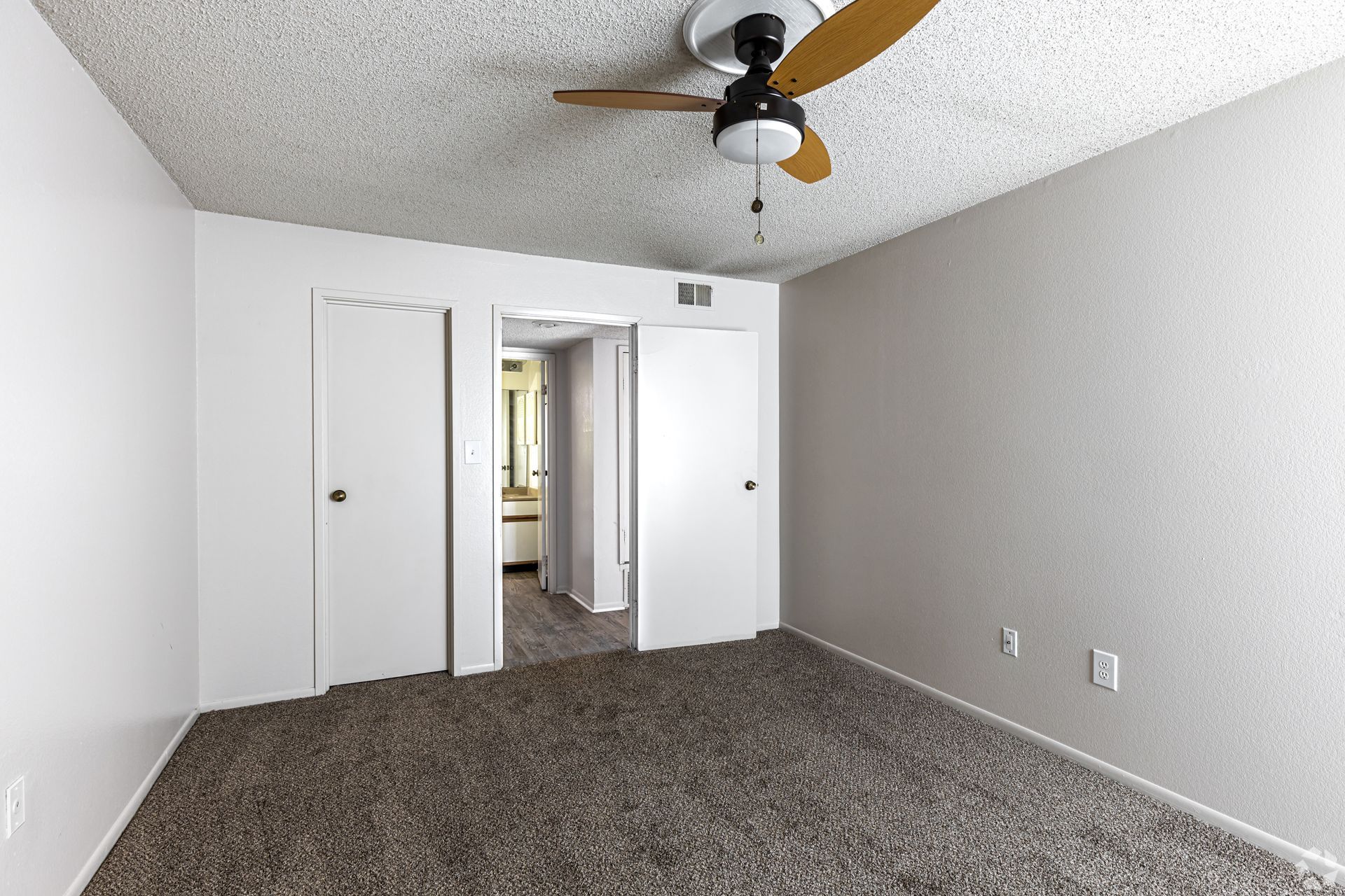 Empty bedroom with white walls, carpet, closet doors, ceiling fan, and open doorway to a bathroom.