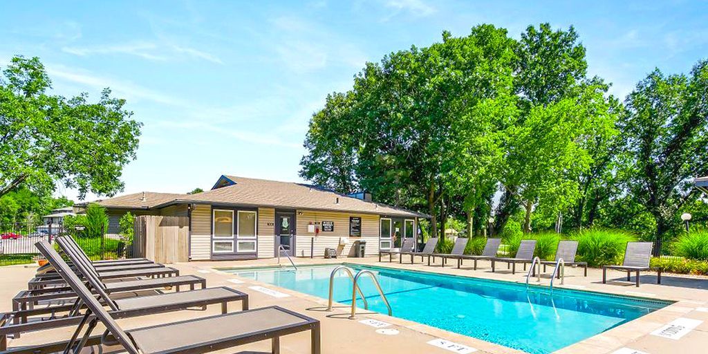 Pool area with lounge chairs, building, and trees on a sunny day.