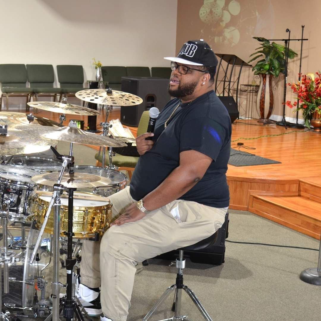 A man is sitting in front of a drum set wearing a hat with the number 308 on it
