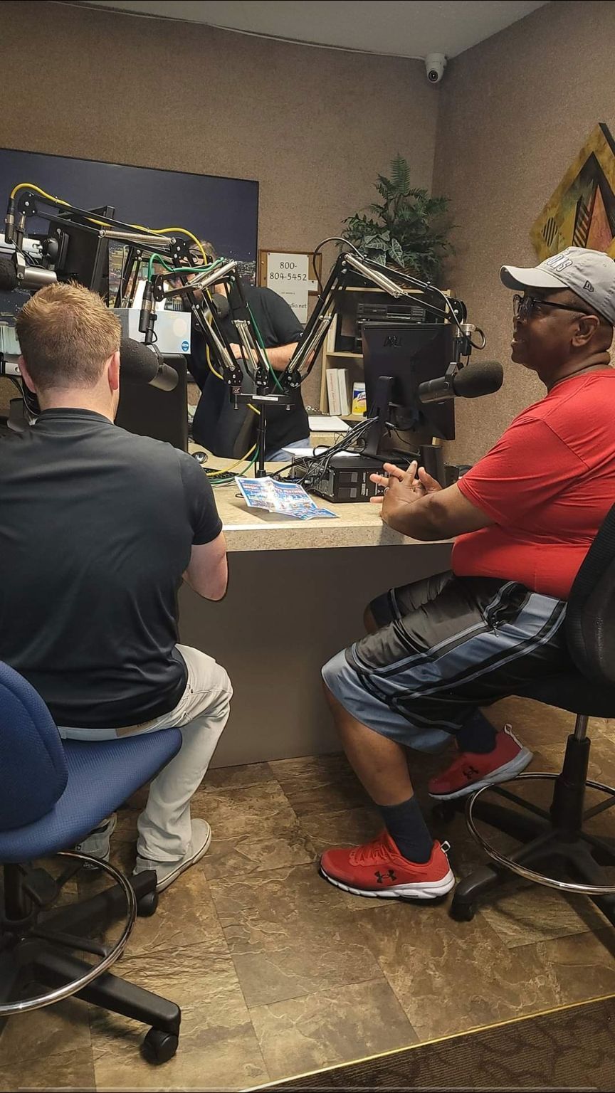 A group of men are sitting at a table in front of microphones in a radio studio.