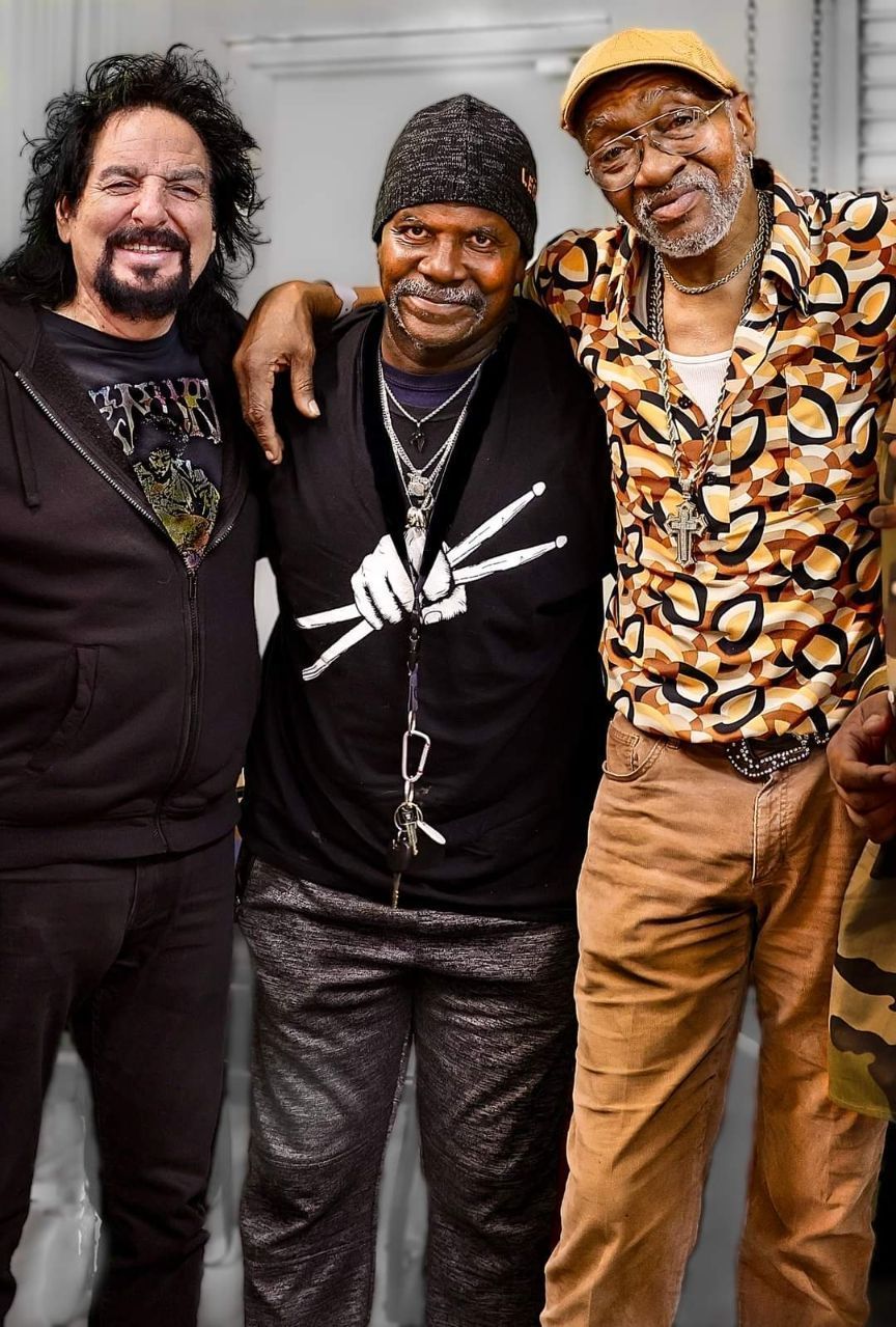 Three men are posing for a picture with one wearing a black shirt with a hand on it