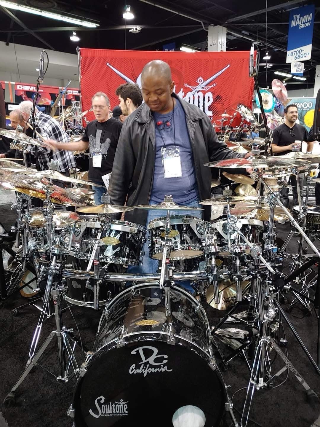 Man with a bald head in a leather jacket stands behind a large drum set at an event with other people.