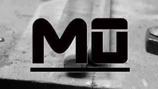 Black "MU" logo with a line underneath against a blurred industrial background.