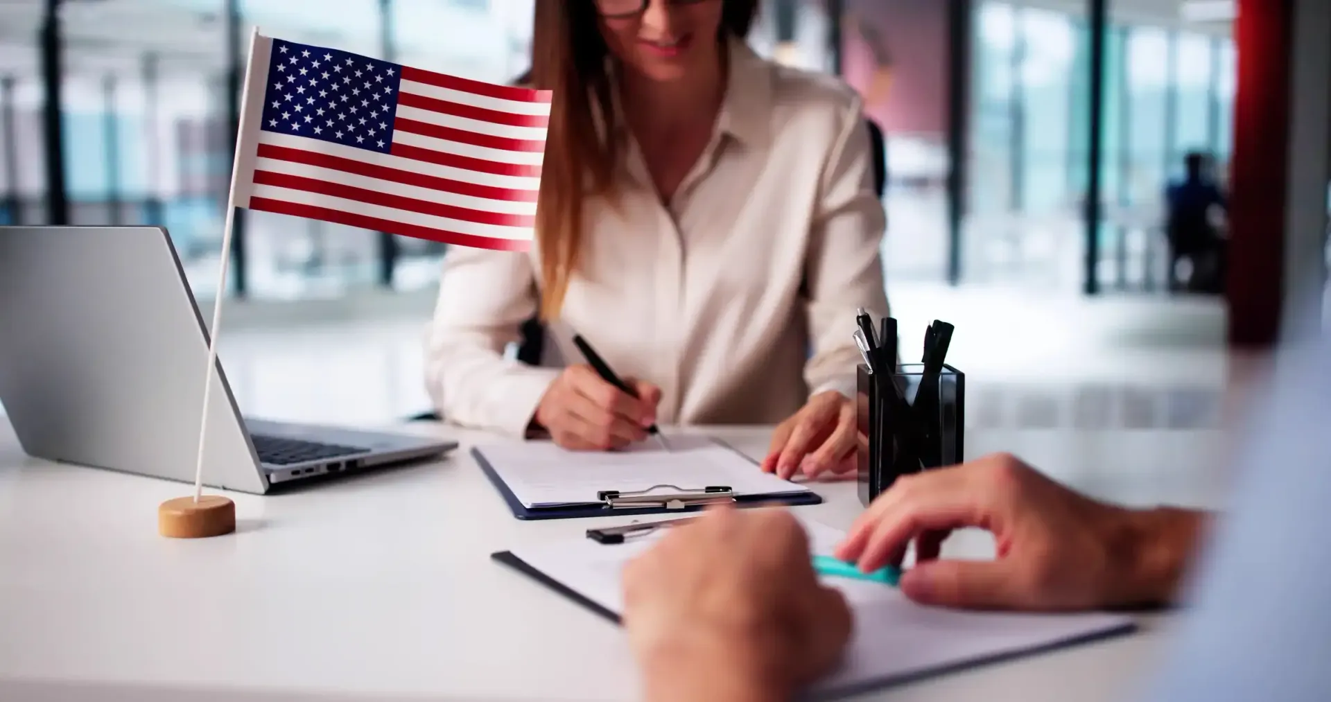Person signing paperwork with another person at a desk, US flag, laptop, pens.