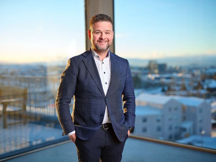 A smiling professional in a navy blazer stands with hands in pockets against a backdrop of a city view through windows.