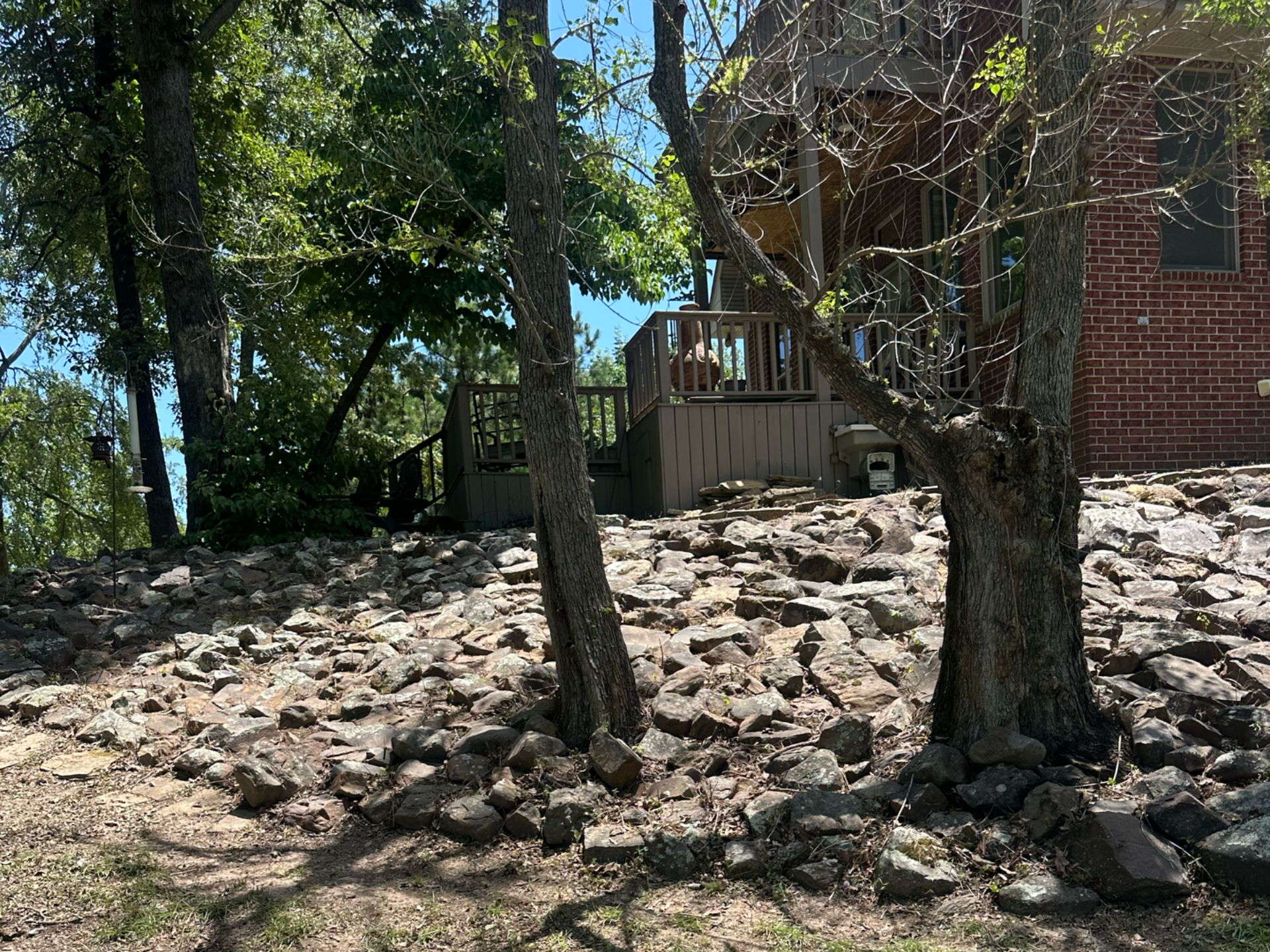 Rocky hillside with trees in front of a wooden deck and brick building on a sunny day.