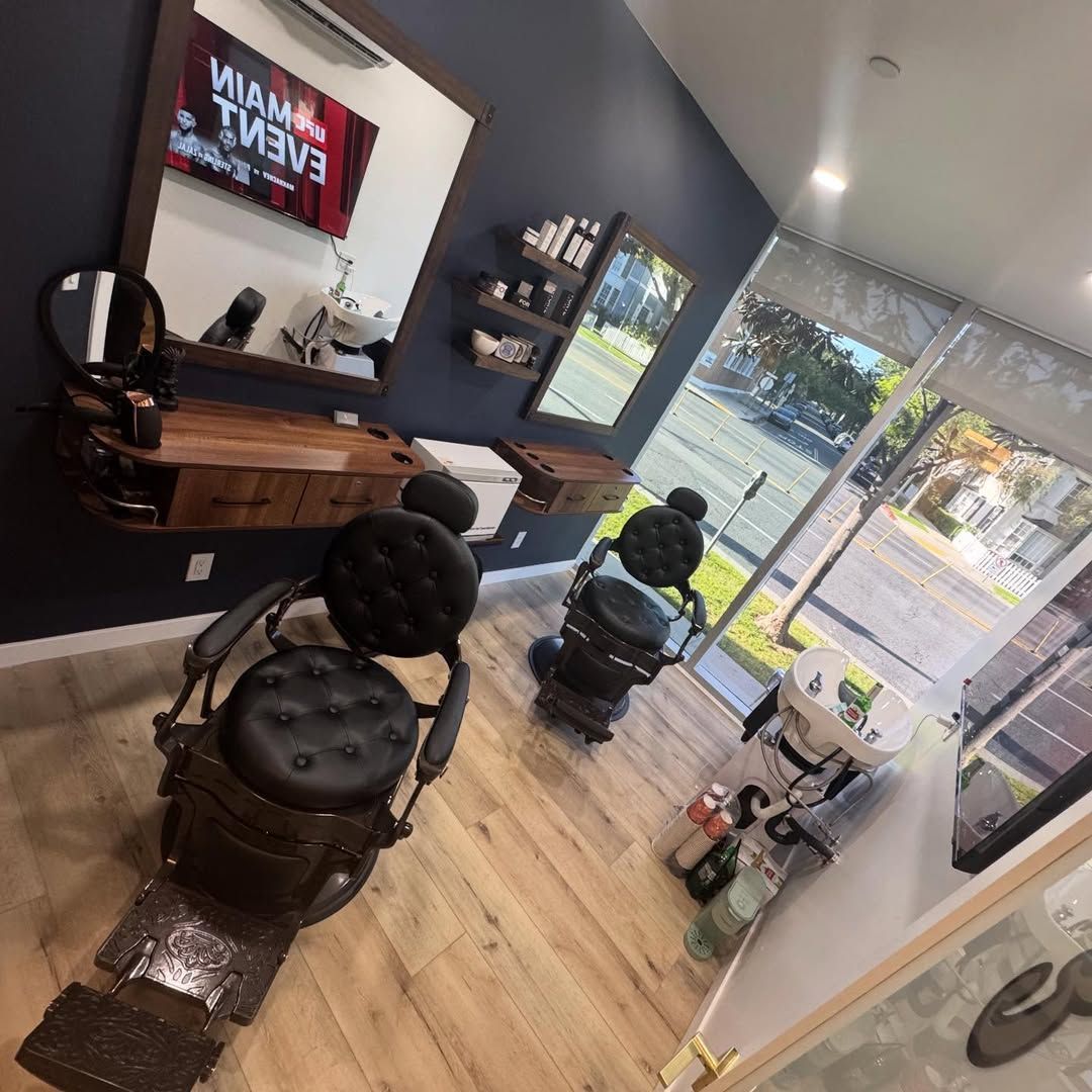 Barbershop interior with two black chairs, wall mirror, and sunlight through the glass door.