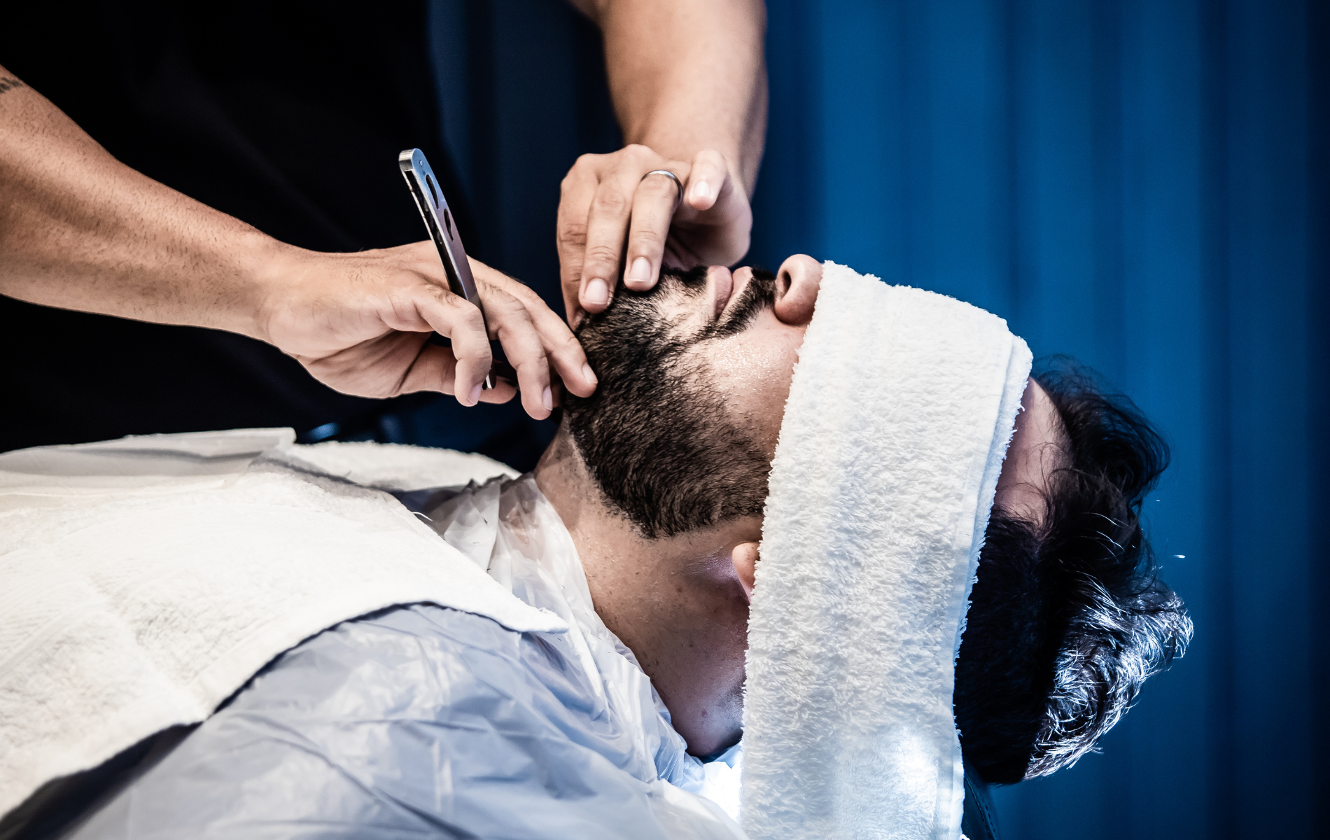 Barber straightens a man's beard with a comb and clipper, with a towel around his head.