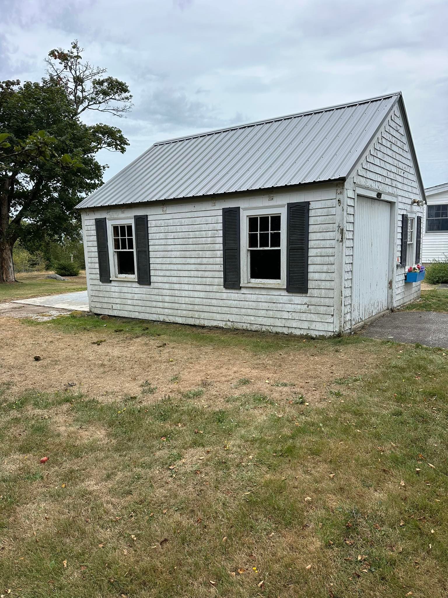 White shed with dark shutters, metal roof, on a grassy plot under a cloudy sky.