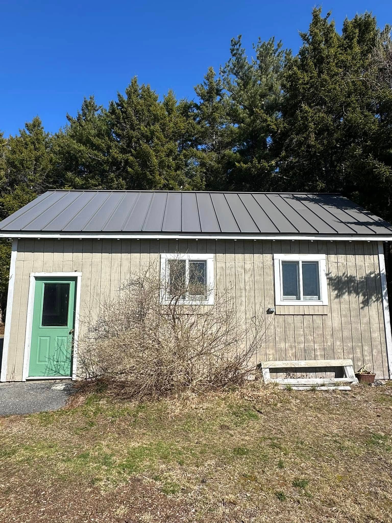 Small, gray, shingled building with green door and windows, gray metal roof, backed by trees, set on a patch of grass.