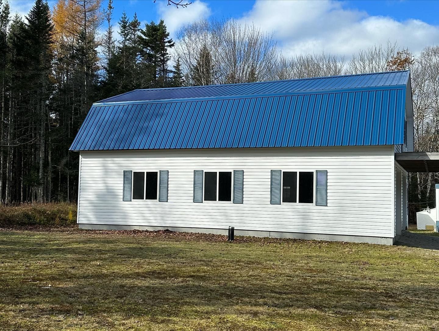 White building with a blue corrugated metal roof. Three windows with shutters, green lawn, trees in background.