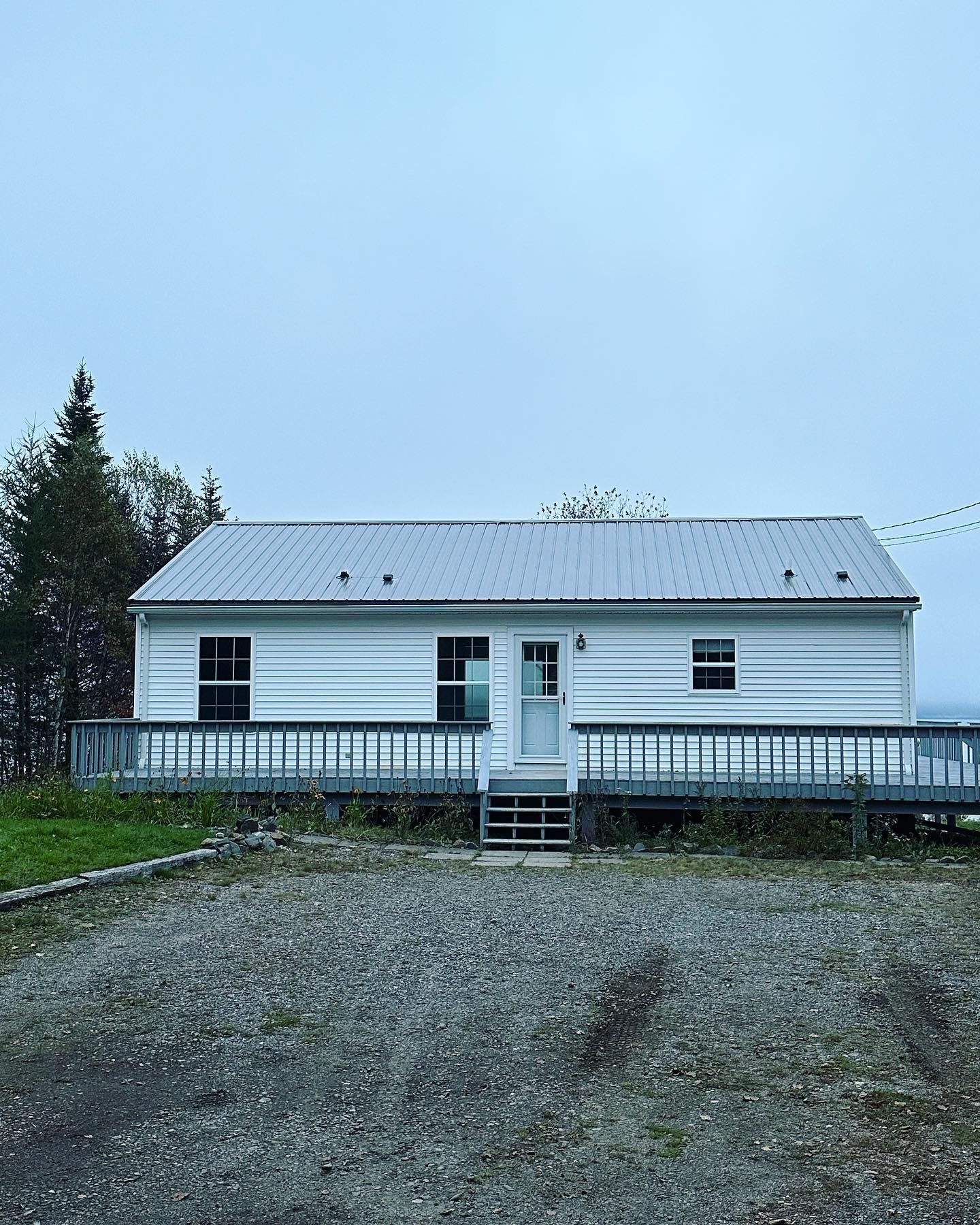 White cabin with a metal roof and a porch on a gravel lot under a cloudy sky.