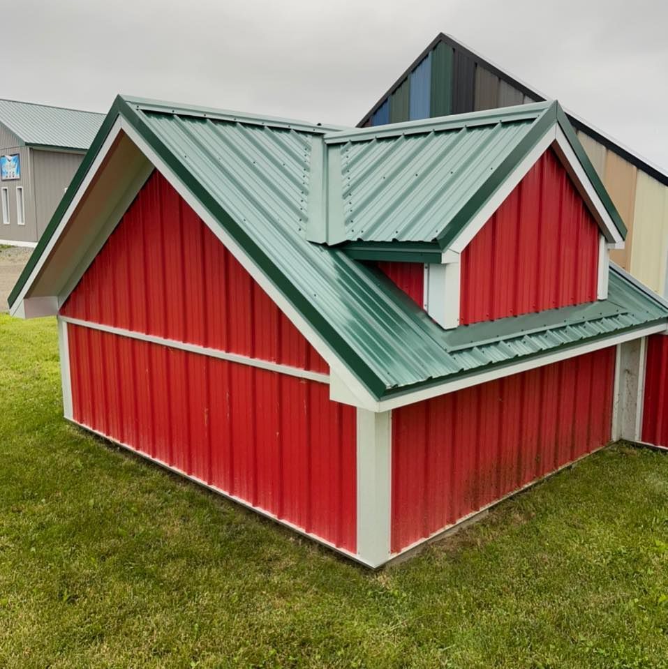 Red barn-like structure with green metal roof and white trim, sitting on grass, with other structures in background.