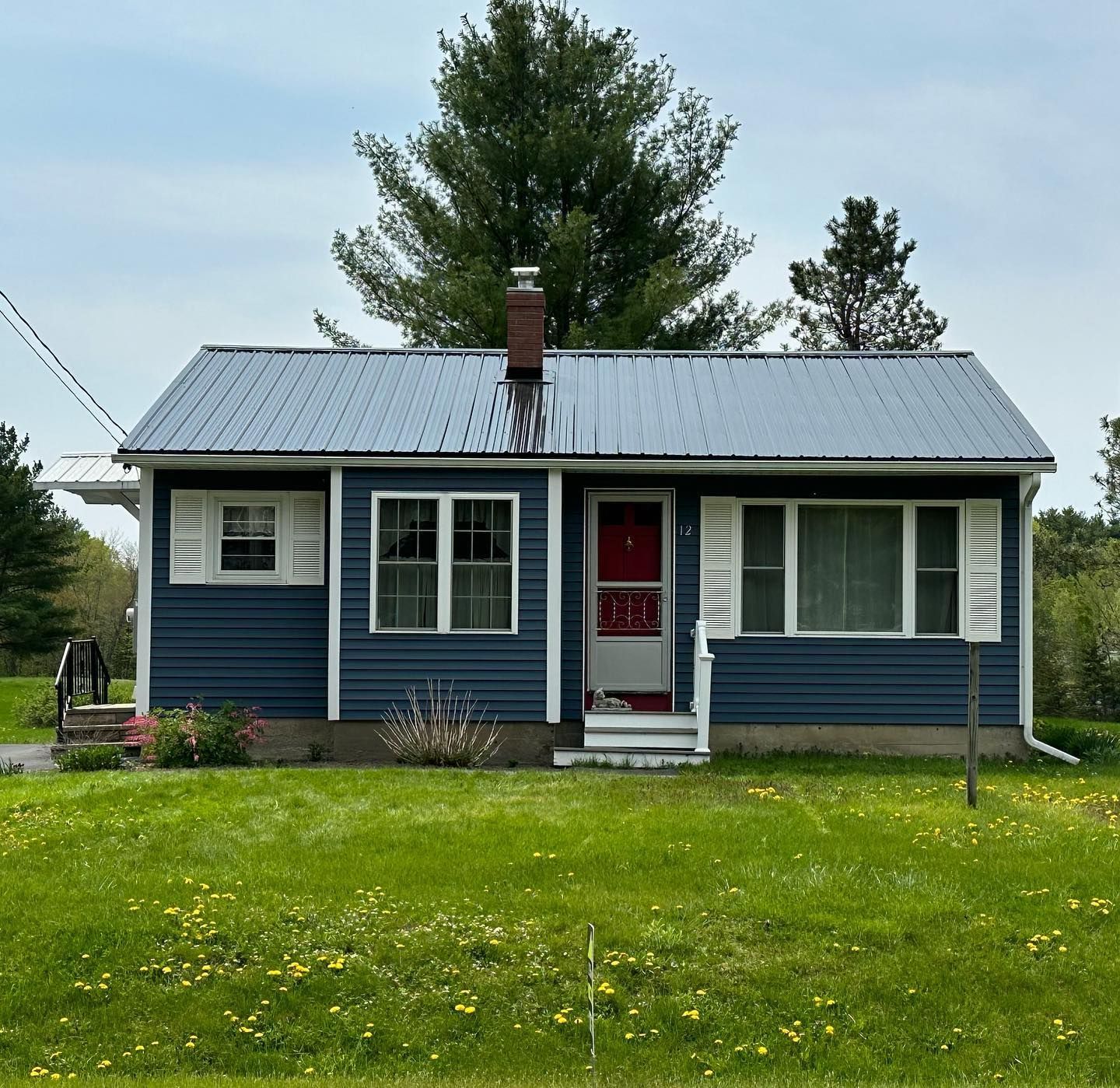 Blue cottage with white trim, metal roof, chimney, and shutters. Green lawn with spring flowers.