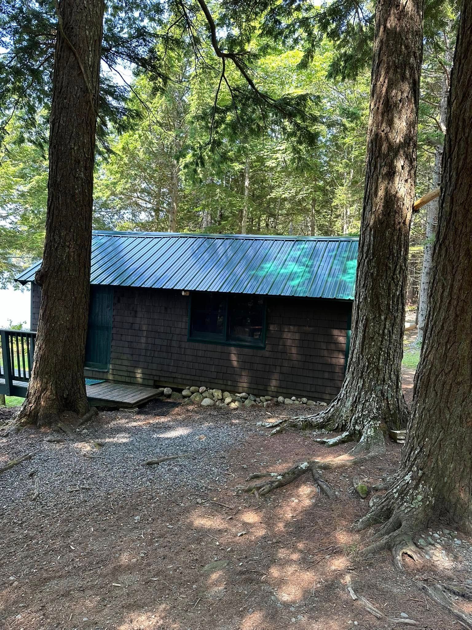 Small cabin with a dark brick facade and metal roof, framed by trees in a wooded area.
