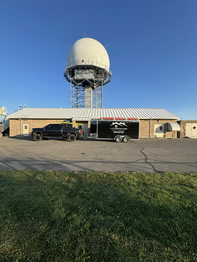 Black truck and trailer parked in front of a white dome radar tower and a low building under a blue sky.