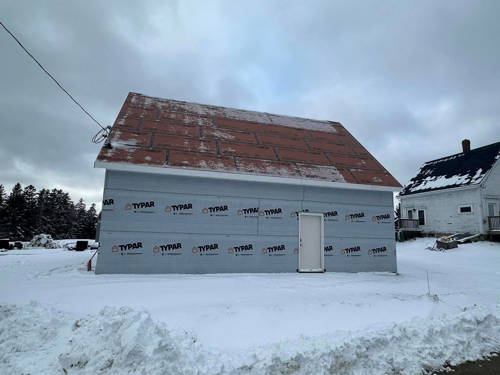 A building under construction covered in Tyvek, with a partially installed brown roof, in a snowy setting.